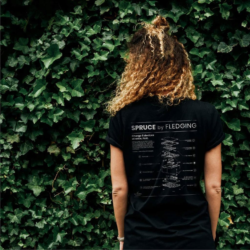 Woman turned around, showing the back of black Spruce t-shirt with an exploded graphic of Spruce Charger's components. Green foliage for background.